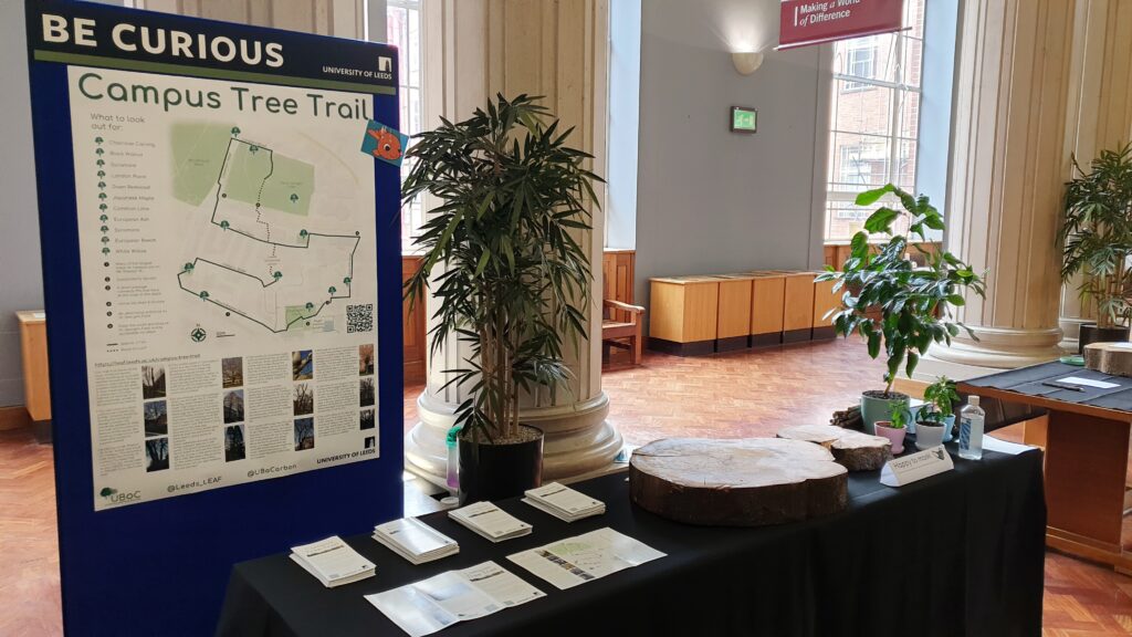 Stall with leaflets, seedlings and a log on top. A poster showing the tree trail is behind.