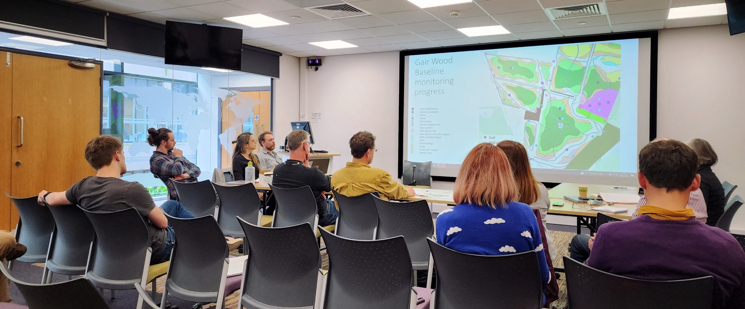 A seminar room with 10 academics in discussion around a powerpoint slide entitled 'Gair Wood baseline monitoring progress'. The slide also shows a map of the site but details are too small to be legible.
